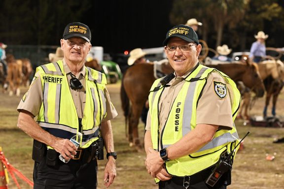 Two elderly white men wearing tan and black sheriff's posse uniforms and bright yellow safety vests