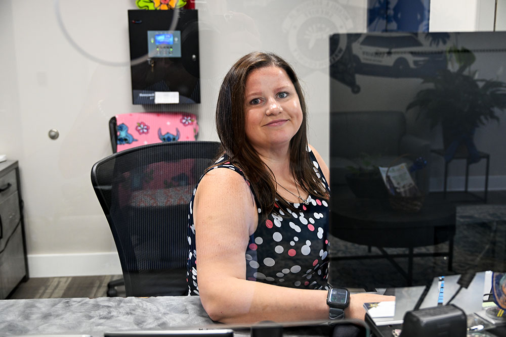 White female with dark brown hair sitting at a desk behind a computer screen smiling