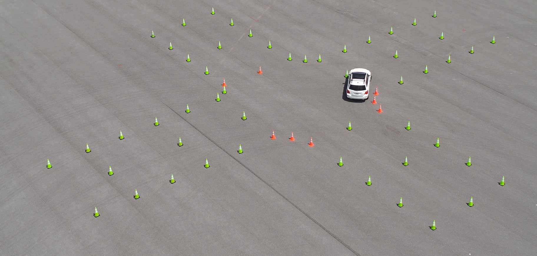 A white car navigates through an empty paved area marked with green and orange traffic cones arranged in a pattern for Teen Driver Challenge.
