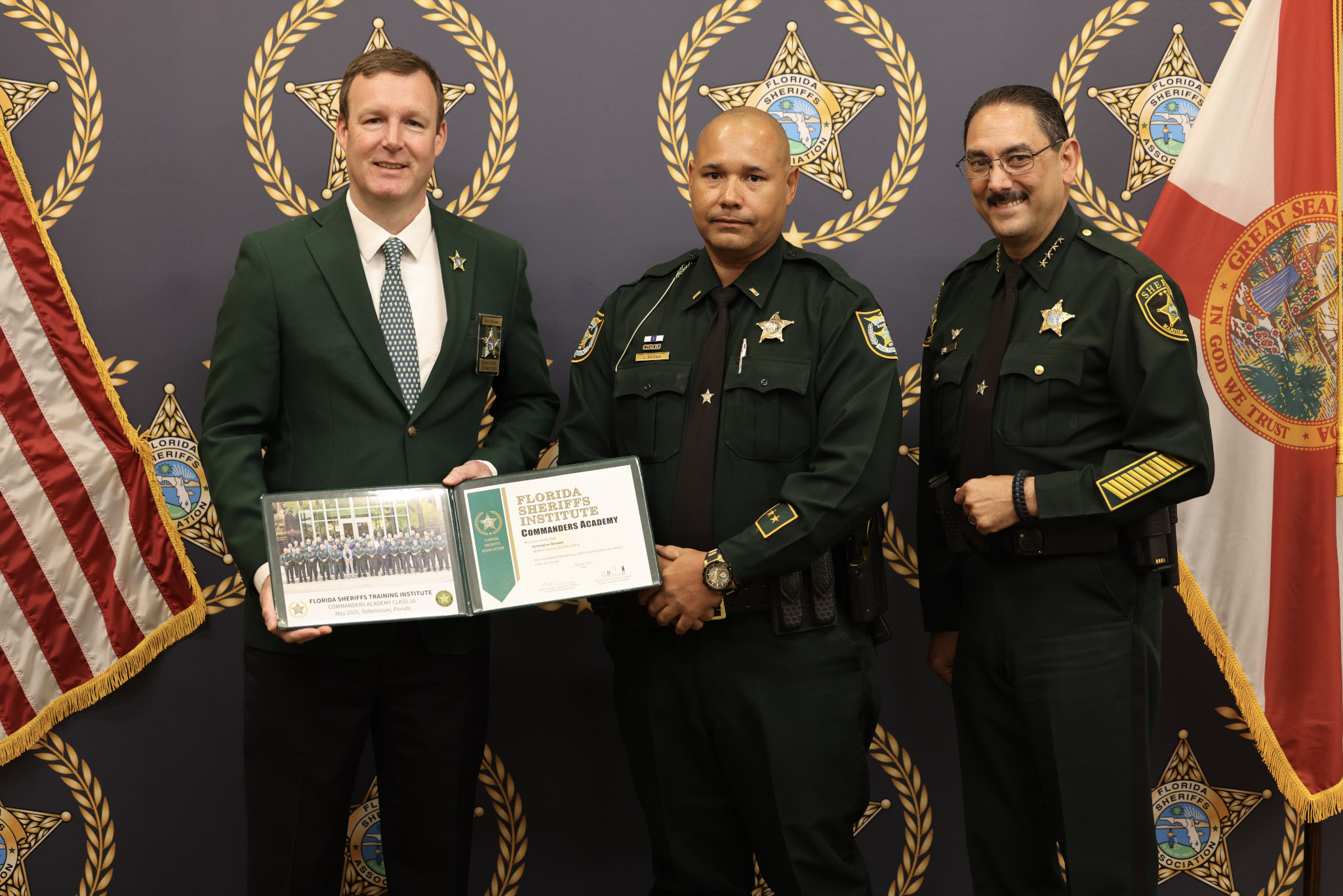 Three men stand in front of a Florida Sheriffs Association backdrop, posing for a formal photo. The man in the center, dressed in a green sheriff’s uniform, holds a certificate from the Florida Sheriffs Institute Commanders Academy along with a class photo. He is flanked by two other men—one on the left in a green blazer with a badge and the other on the right in a green sheriff’s uniform with sergeant stripes. Both American and Florida state flags are displayed on either side of the group, reinforcing the official and ceremonial nature of the occasion.