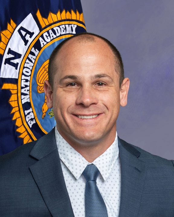 White male with short dark brown hair wearing a blue suit and tie standing in front of an FBI National Academy flag