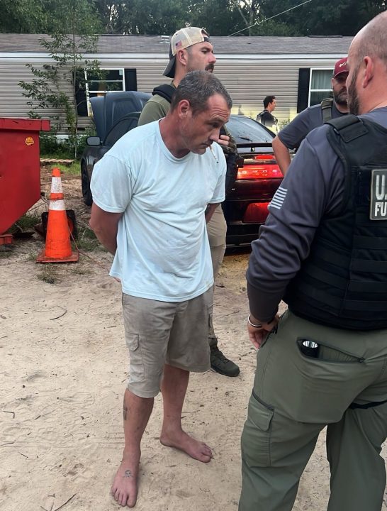White male wearing khaki shorts and a white shirt with a cigarette in his mouth surrounded by law enforcement officers