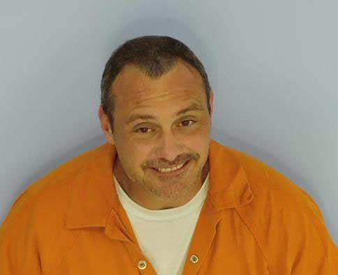 White male with short brown hair smiling wearing an orange jail jumpsuit