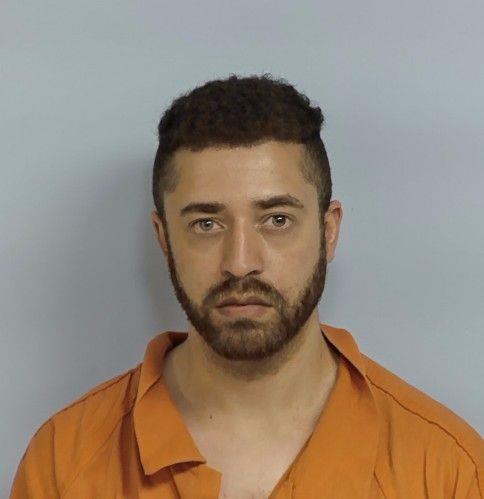 Mugshot of a man wearing an orange prison jumpsuit, standing against a plain light gray background.