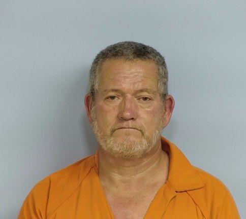 A white male with white and gray hair and goatee wearing an orange jumpsuit standing in front of a light blue wall.