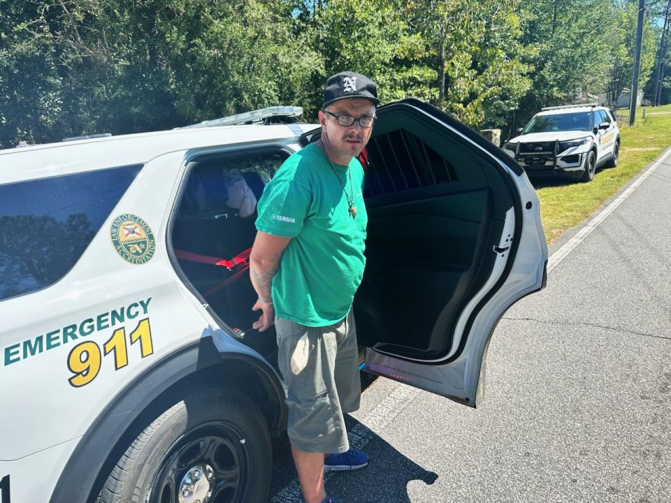 White male wearing a green shirt and black hat with his hands cuffed behind his back in front of a patrol car