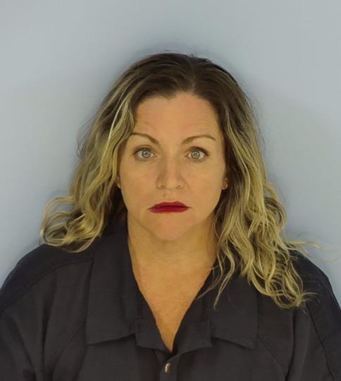 Mugshot of a woman with wavy blonde hair wearing a dark-colored collared shirt, looking directly at the camera against a plain light blue background.
