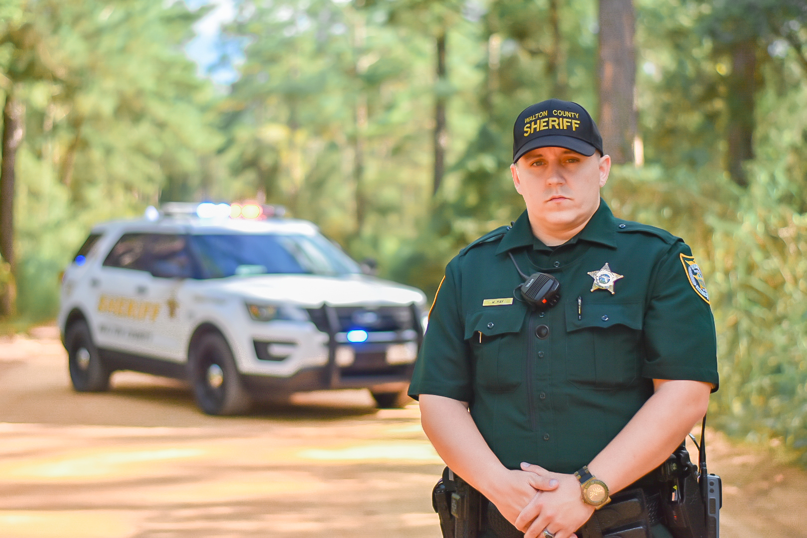 Walton County Sheriff’s Deputy William May in uniform stands on a wooded road with a patrol vehicle behind him, emergency lights flashing.