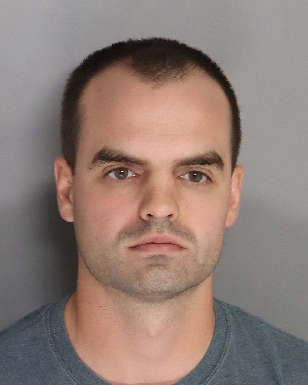 Mugshot of a man with short dark hair, light skin, and a neutral expression, wearing a gray t-shirt, against a plain gray background.