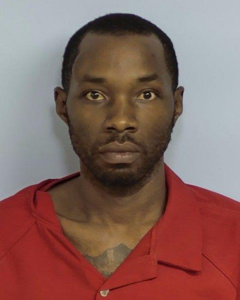 Mugshot of an adult man with short hair and a trimmed beard, wearing a red collared jail uniform, standing against a plain light-blue background.