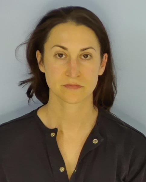 Mug shot of a white female with dark hair wearing a navy blue jumpsuit standing in front of a light blue wall.