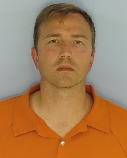 Mug shot of a white male with blue eyes and brown hair wearing an orange jumpsuit standing in front of a light blue wall.