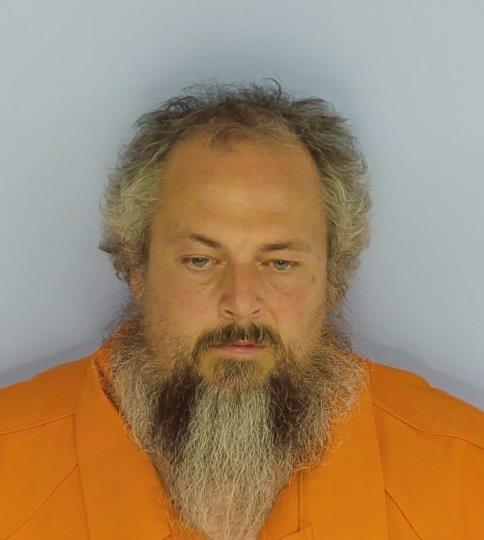 Mug shot of a white male looking down with gray and black hair and a long gray and black beard wearing an orange jumpsuit standing in front of a light blue wall.