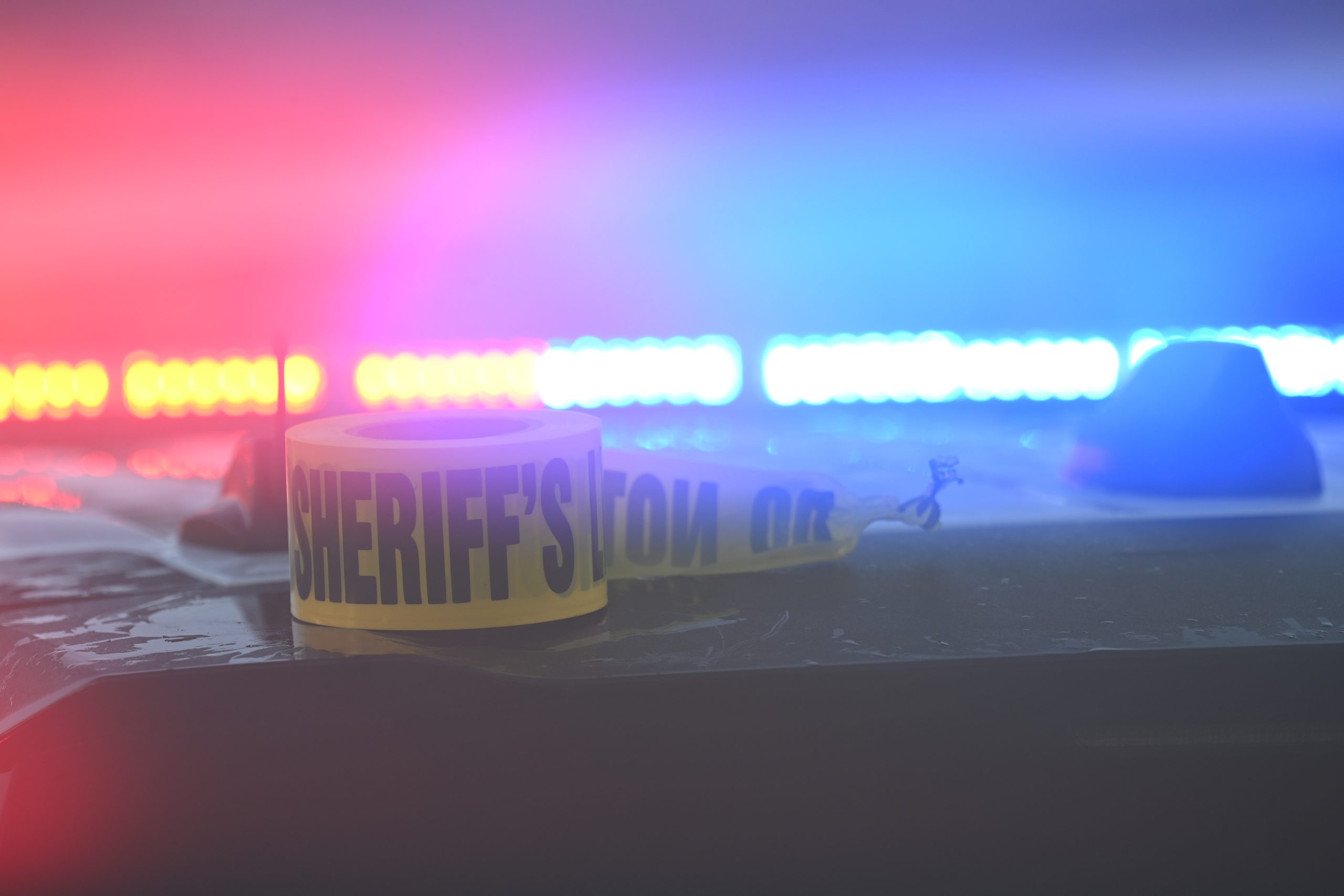 A roll of yellow sheriff’s office caution tape sits on a wet surface, illuminated by the red and blue flashing lights of a police vehicle in the background.
