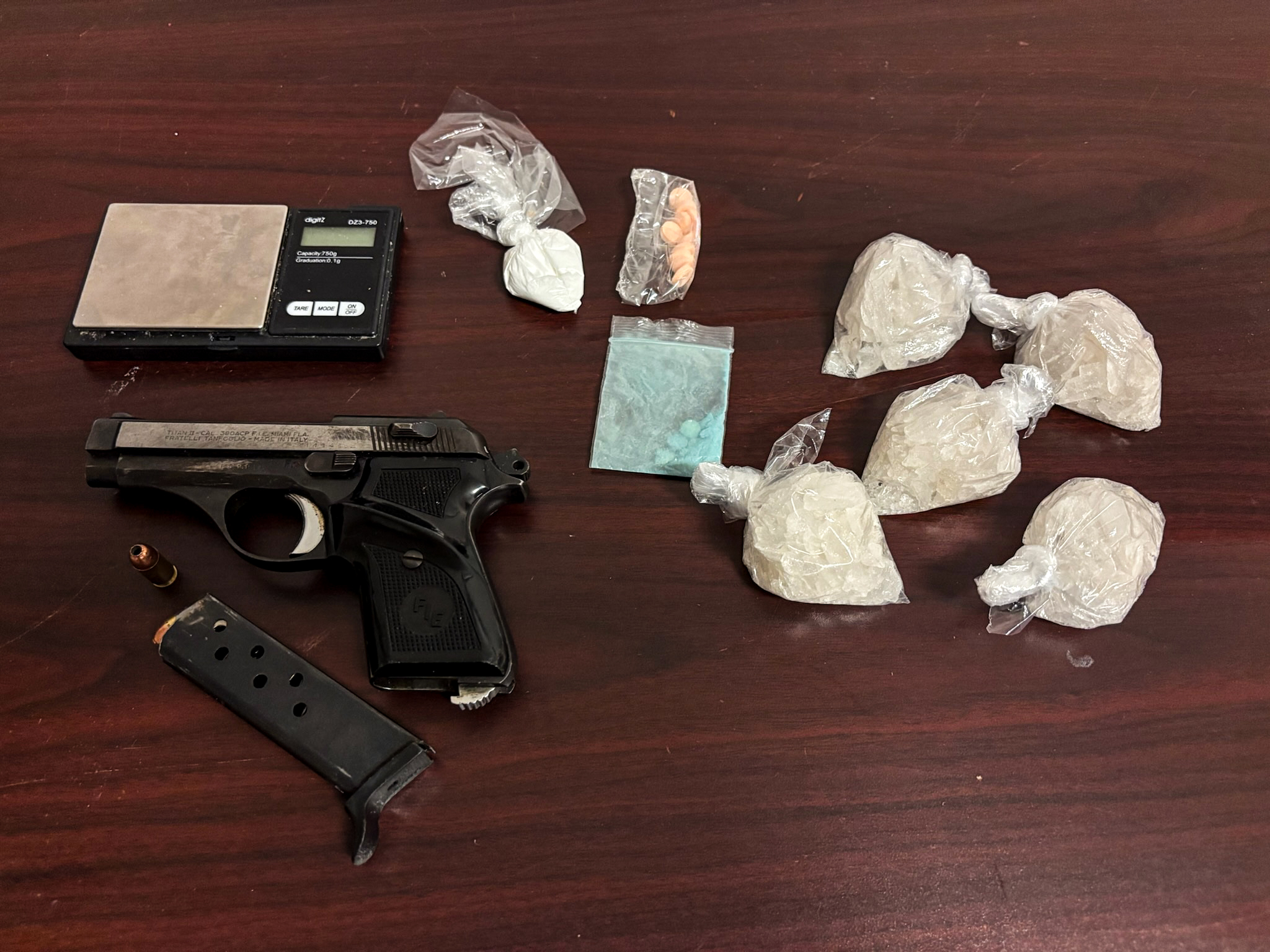 A wood table with a gun and scale and multiple clear plastic bags of white powdery substance and one clear ziploc back with blue pills inside