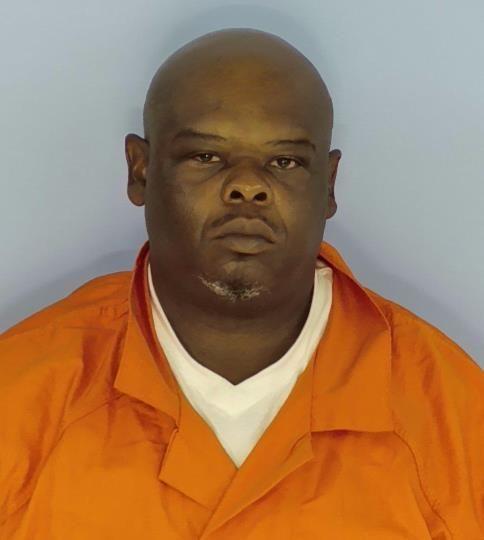 A mug shot of a black man in an orange jump suit with a gray and black goatee standing in front of a light blue background.