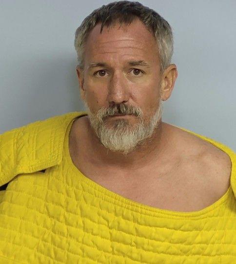 A middle-aged man with short gray hair and a gray beard wearing a yellow quilted safety smock, photographed against a plain backdrop in a mugshot-style image.