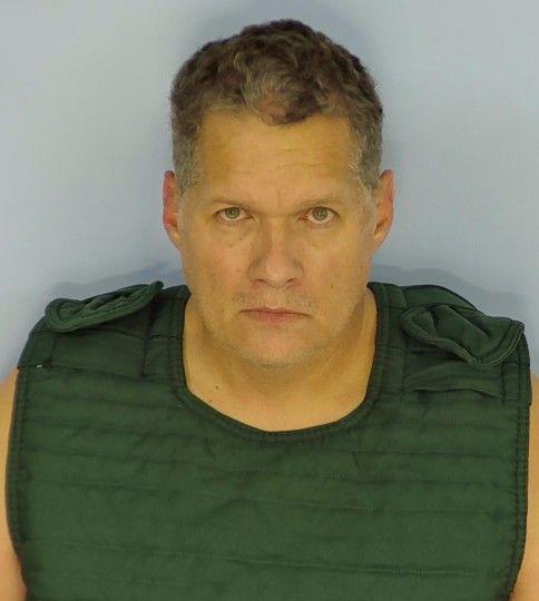 A white male wearing a hunter green orange jumpsuit standing in front of a light blue wall.