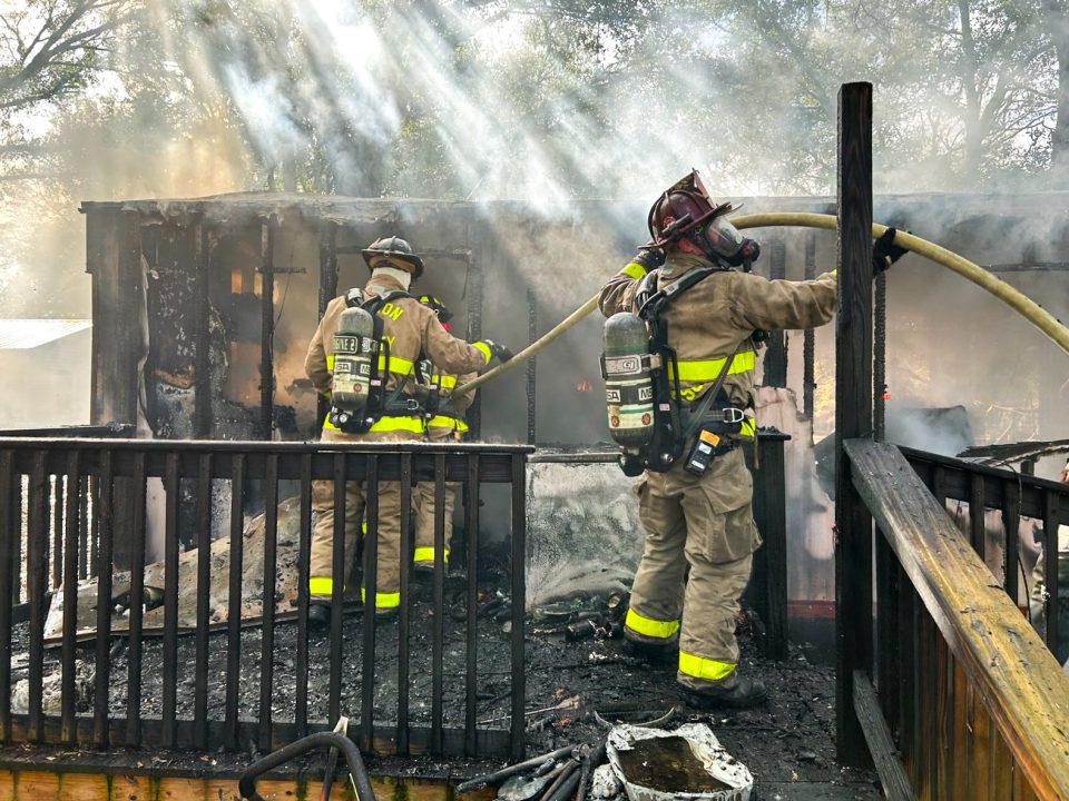 Two firefighters carrying a yellow fire hose on the deck of a burned-down mobile home