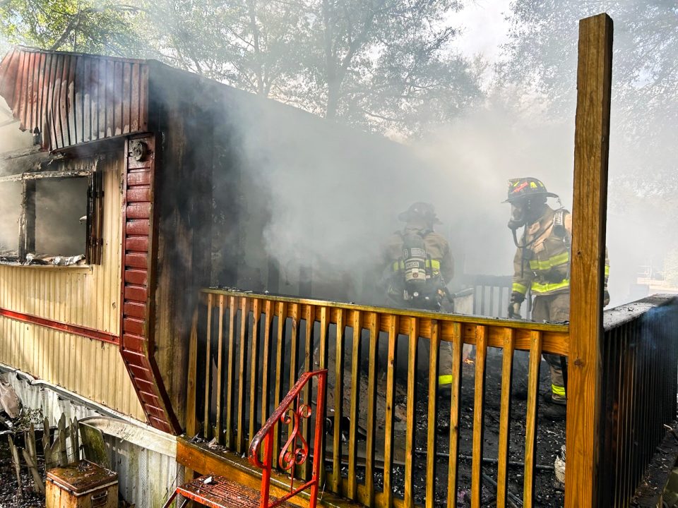 Firefighters on a large back deck behind a mobile home with heavy smoke coming out of it