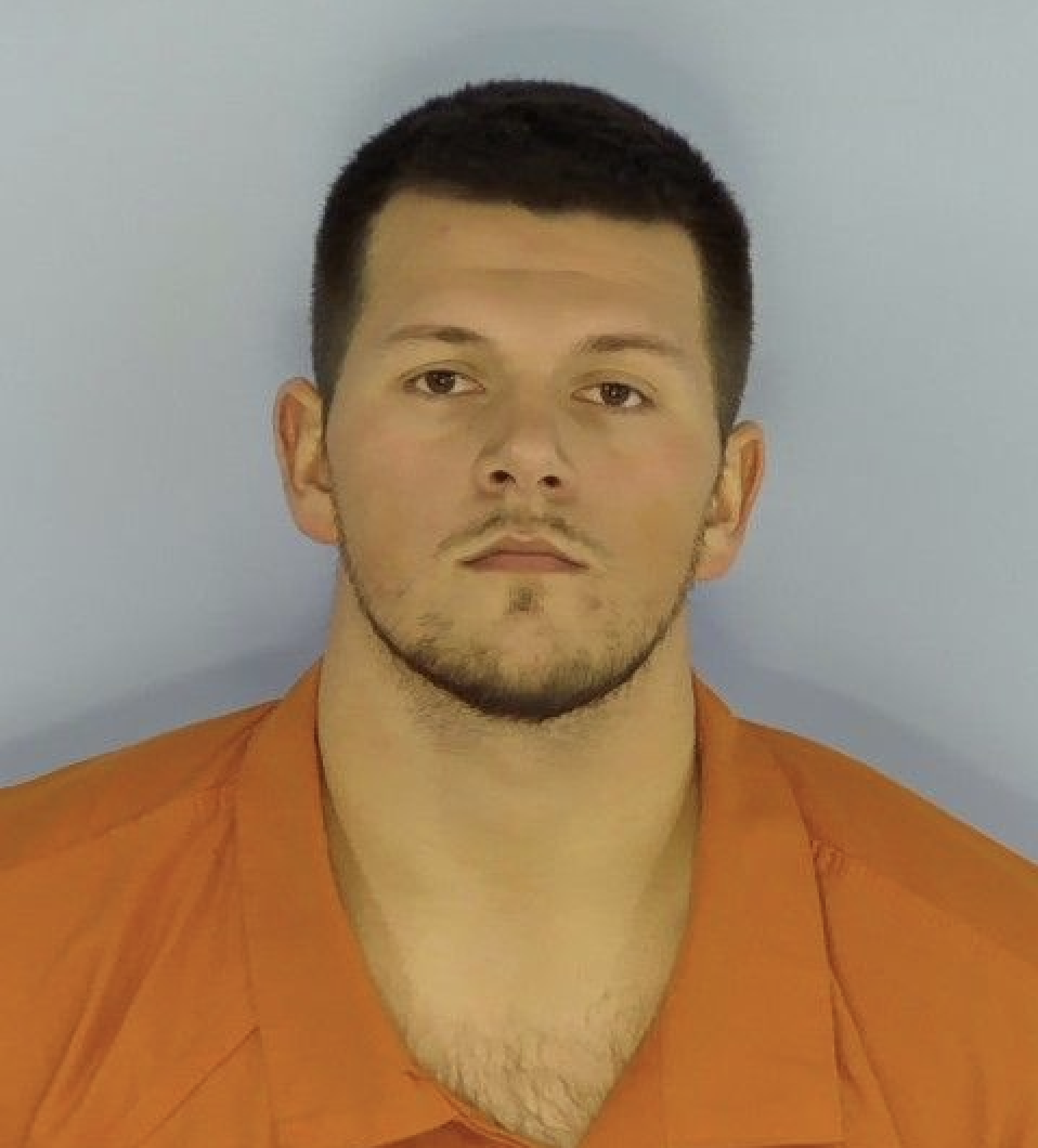 A white male wearing an orange jumpsuit standing in front of a light blue wall.