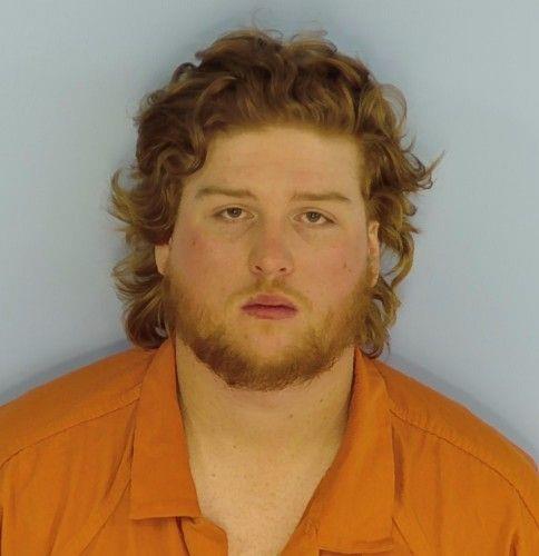 White male with red hair and a red beard wearing an orange jumpsuit standing in front of a light blue wall.