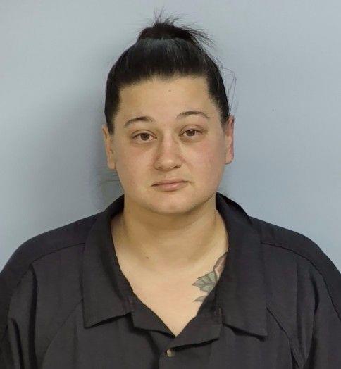 Mug shot of a white woman with dark brown hair wearing a navy jumpsuit standing in front of a light blue wall. 