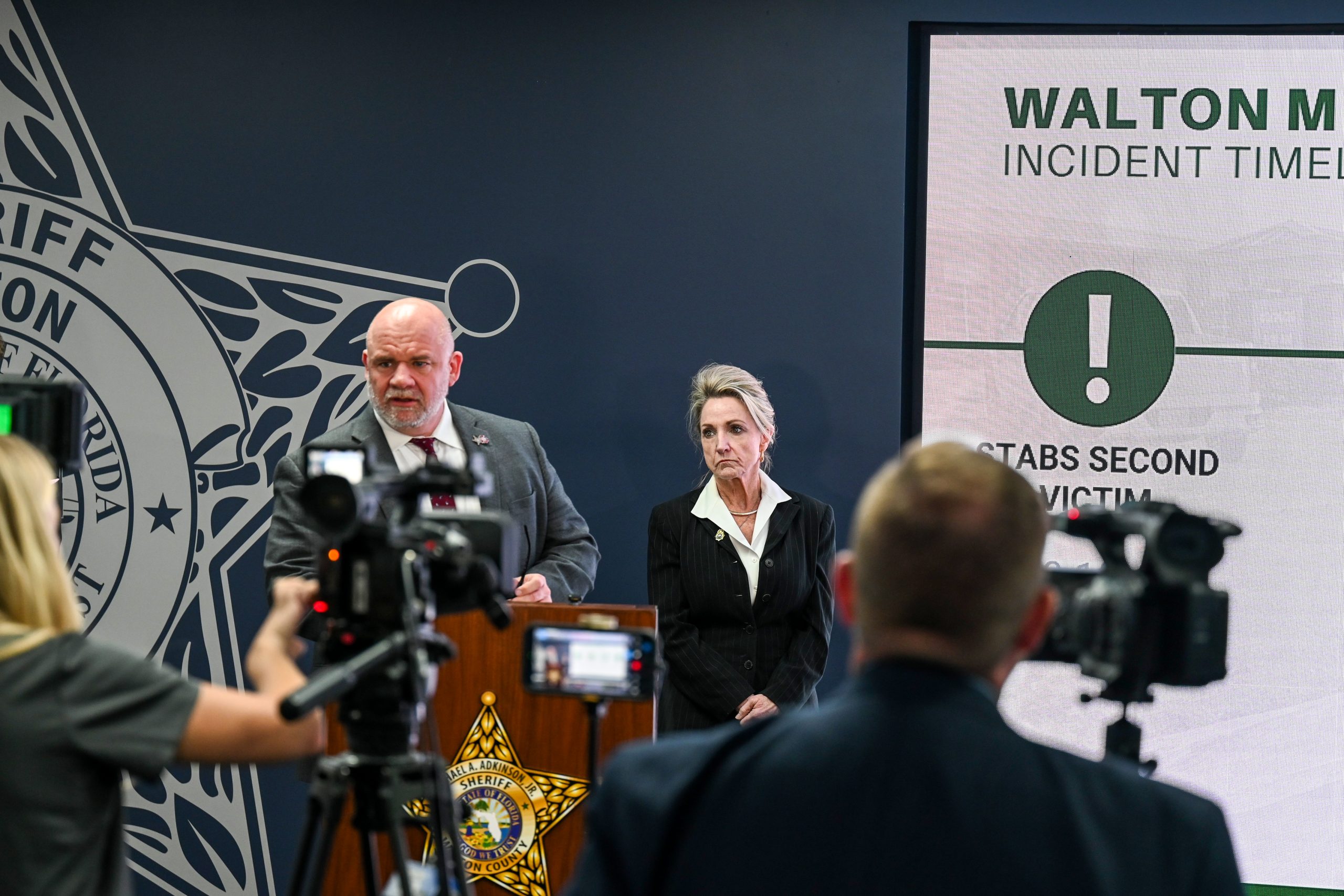 Press conference scene inside a sheriff’s office briefing room. Two individuals stand at a podium with the sheriff's star emblem on the front, surrounded by multiple video cameras and media personnel recording the event. A large display screen to the right shows the heading ‘Walton M… Incident Time…’ along with a warning symbol and partially visible text. The background wall features a large sheriff’s office insignia.
