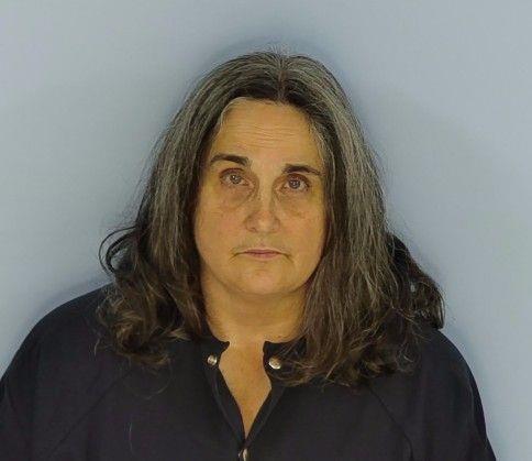 Mug shot of a white female with graying dark hair standing in front of a light blue wall wearing a navy blue jump suit.