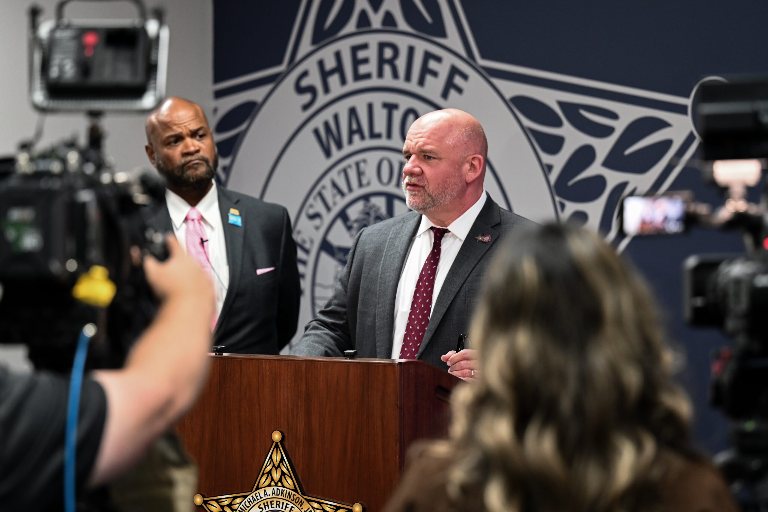"Press conference setting inside a sheriff’s office briefing room. Two individuals stand behind a wooden podium displaying a sheriff’s star emblem. Several cameras and media personnel are positioned in the foreground recording the event. The background features a large wall graphic of a sheriff’s office badge with the text ‘Sheriff’ and ‘Walton’ visible.