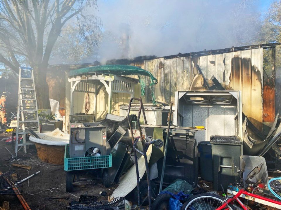 Light colored single-wide mobile home with a lot of junk in the yard and smoke coming from the home