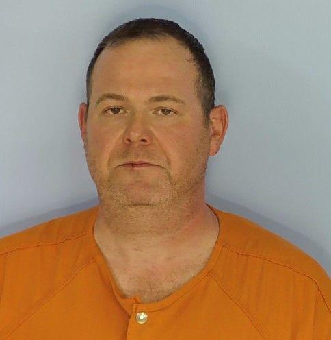 An individual wearing an orange button‑front garment is photographed against a plain light‑colored background. The person is centered in the frame, shown from the upper chest to the top of the head.