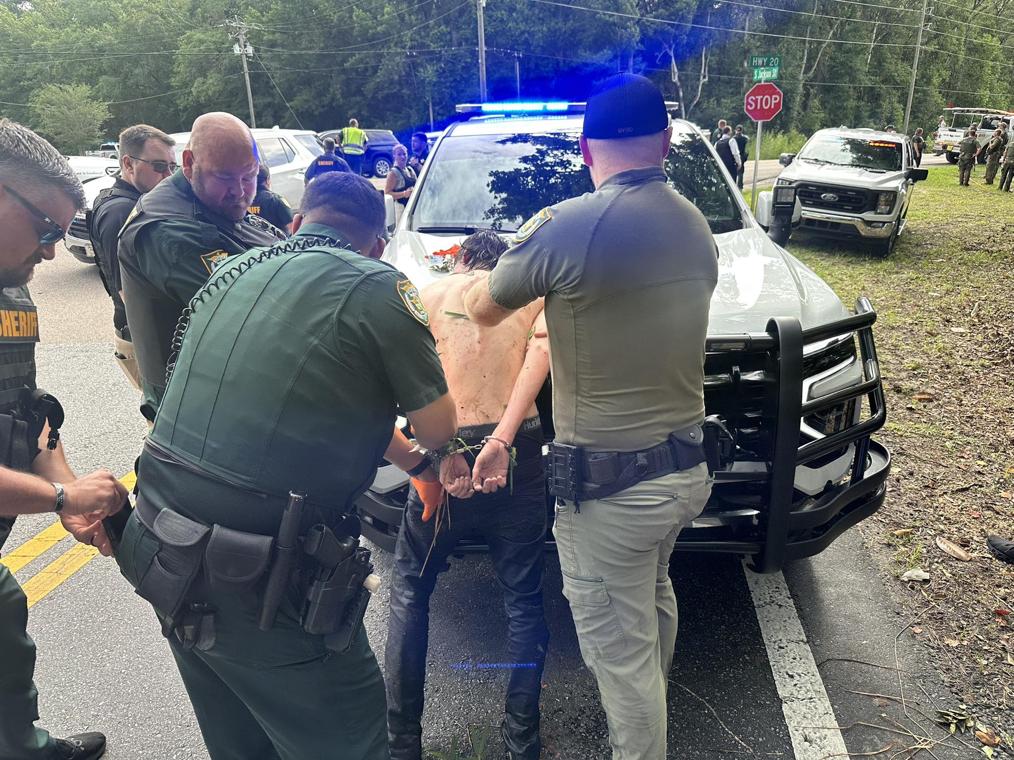 Several uniformed individuals stand on a roadway near a marked vehicle with flashing lights while securing the hands of another individual whose hands are restrained behind the back. Additional vehicles, personnel, and roadside signs are visible in the background along a tree‑lined area.