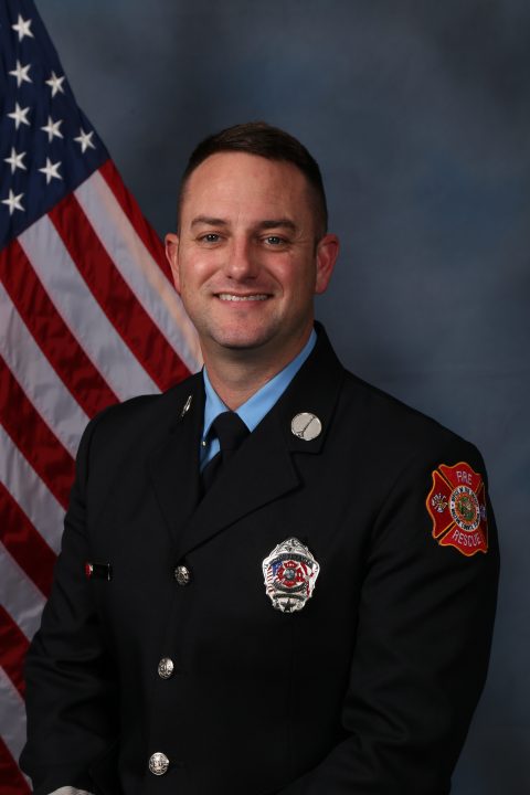 White male firefighter with dark brown hair wearing a navy blue Class A uniform and a light blue shirt