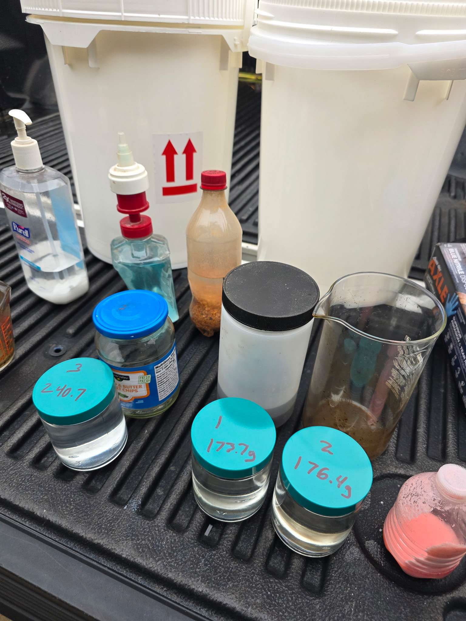 A collection of assorted containers and chemicals is arranged on the bed of a pickup truck. The items include large white buckets, plastic bottles, jars with blue lids, beakers containing liquids or residue, and various labeled containers with handwritten numbers.