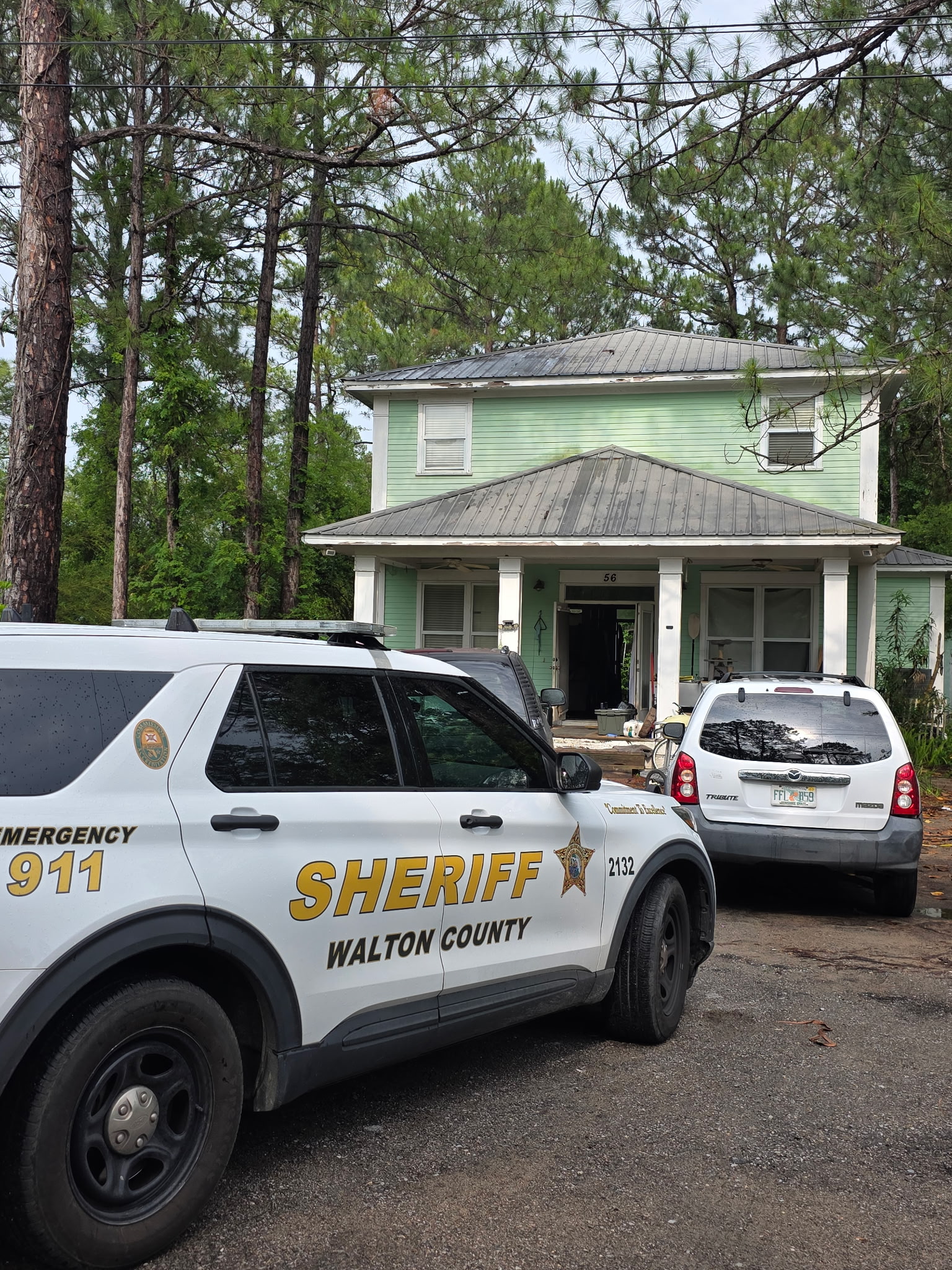 Two marked Walton County Sheriff’s Office vehicles are parked in front of a light green two‑story house surrounded by tall pine trees. The front door of the house is open, and the driveway contains debris and equipment.