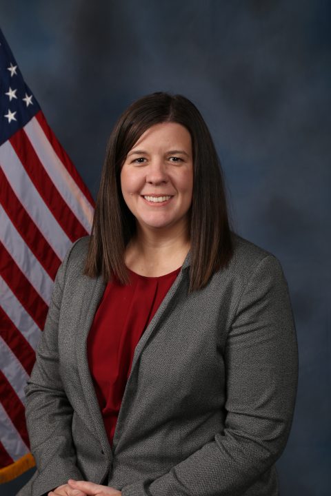 White female with dark brown hair wearing a red shirt and a gray suit jacket