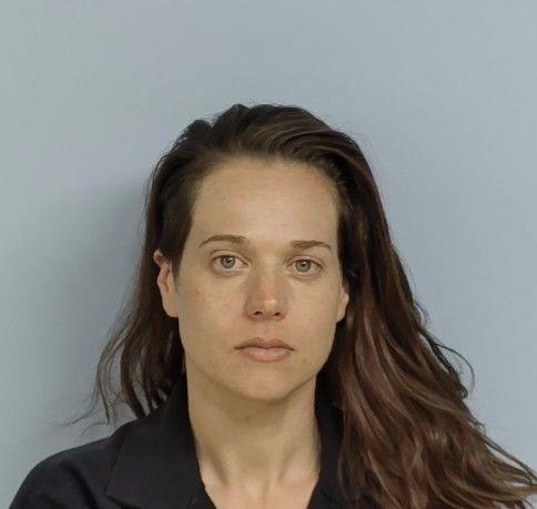 A head‑and‑shoulders photograph of an adult individual facing the camera against a plain light‑blue background. The person has long, dark hair that falls over one shoulder and is wearing a dark collared shirt. The individual’s expression is neutral, and no additional objects or text are visible in the image.