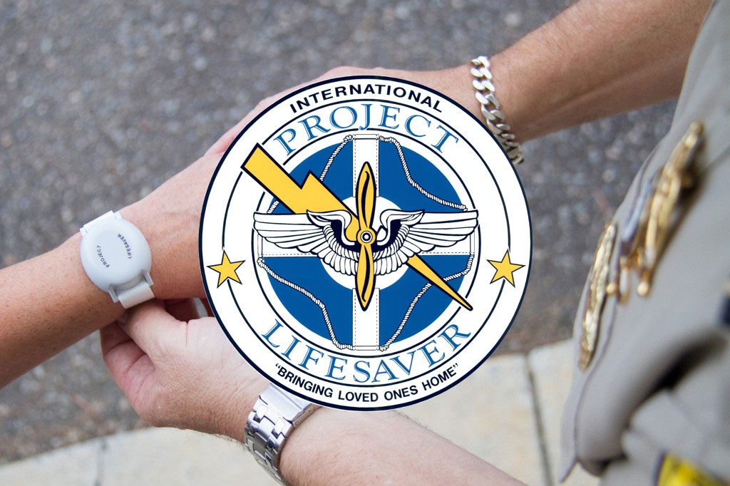 A close-up photo shows two people’s hands. One person is wearing a white Project Lifesaver wrist transmitter. Another person, wearing a uniform with visible pins and a chain bracelet, is gently checking or adjusting the device. The Project Lifesaver logo is centered over the image. The logo features a blue and white circular emblem with a pair of wings, a lightning bolt, and the words “International Project Lifesaver” and “Bringing Loved Ones Home.”